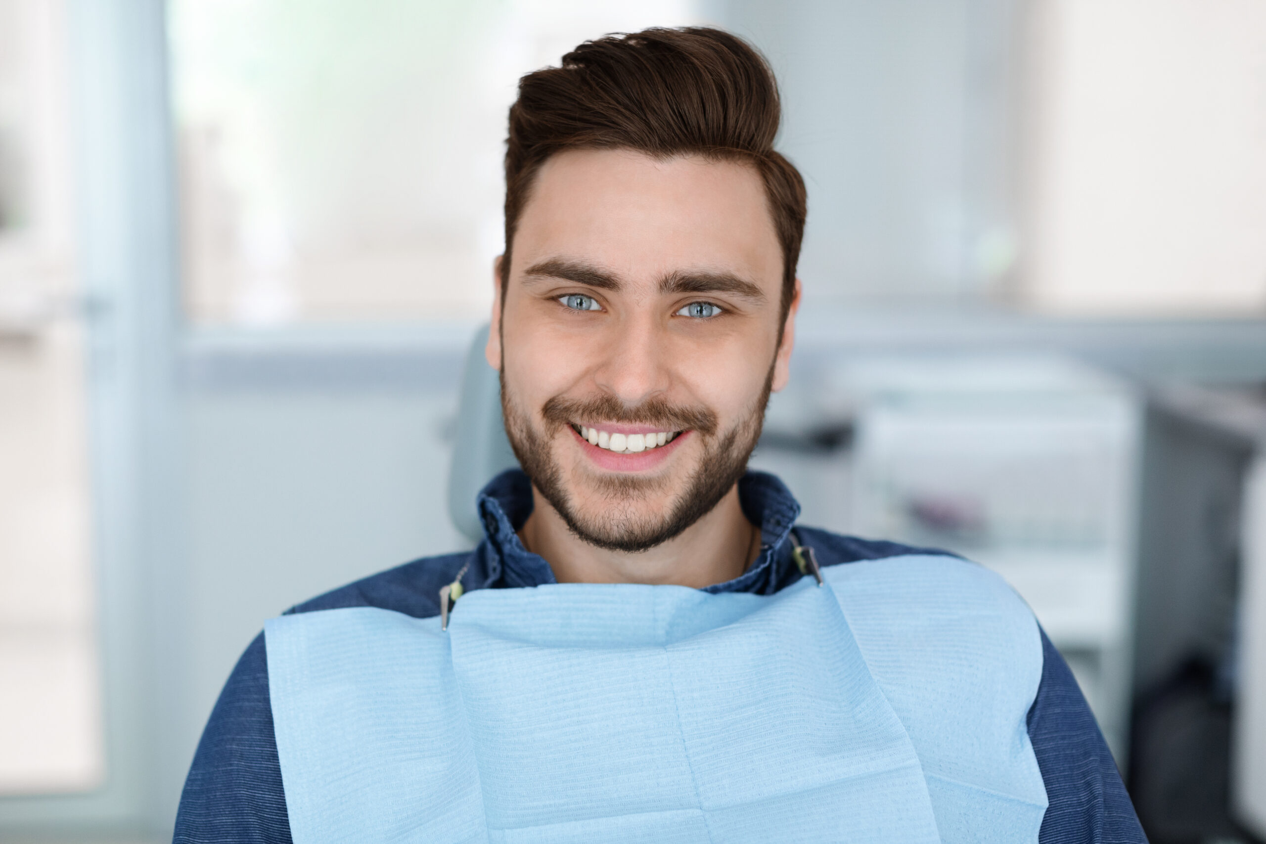 Cheerful bearde man patient sitting in dental chair and smiling at camera, copy space. Positive millennial guy showing his beautiful white smile after treatment at modern dental clinic